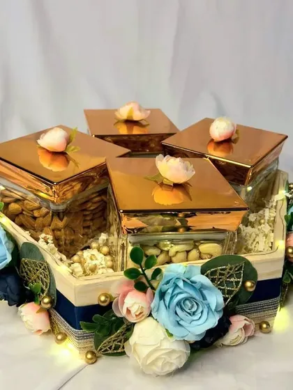 This elegant dry fruit box is a popular choice for trousseau gifting. The square glass jars with rose gold lids are arranged in a floral-decorated wooden tray, combining modern style with a touch of tradition.