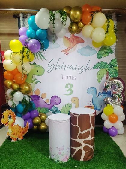 A colorful and fun dinosaur theme for Shivansh's third birthday. The decor includes a custom backdrop with friendly dinosaurs, a multi-colored balloon arch, and a number '3' foil balloon.