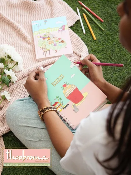 This overhead lifestyle shot for Theobroma merchandise shows their products in a creative, relaxed setting. The composition includes branded notebooks and art supplies on a picnic blanket, suggesting a whimsical and imaginative brand personality.