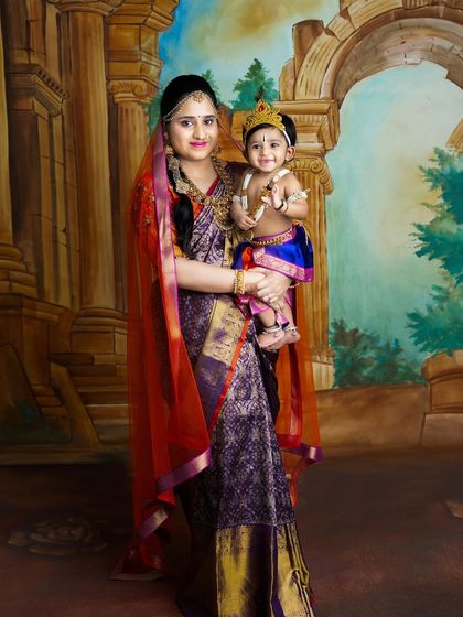 A classic portrait of a mother holding her child, who is dressed as Krishna. The rich colors of their outfits and the painted backdrop create a regal feel.