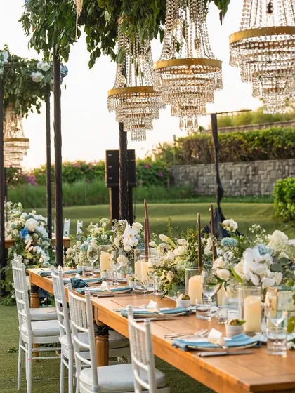 For an elegant outdoor reception, I designed this long dining table setup under a canopy of greenery and crystal chandeliers. This arrangement is perfect for creating a sophisticated and intimate dining experience for your guests.