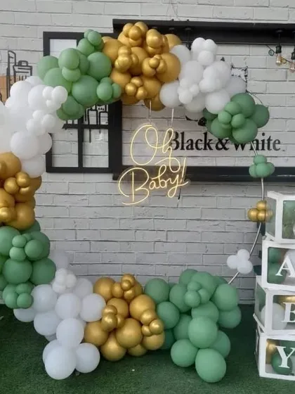 A modern 'Oh Baby' shower setup with a balloon arch in green, gold, and white, and 'BABY' boxes.