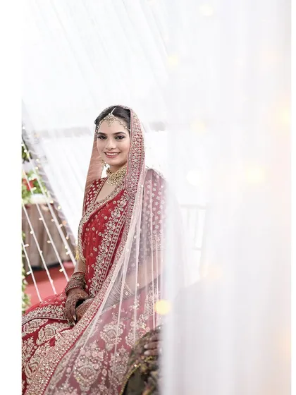 An ethereal portrait of a bride in a stunning red lehenga. The white curtains and soft light create a bright and airy feel, making her the absolute center of attention.