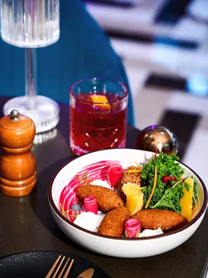 A colorful appetizer bowl served with a red cocktail in a vibrant, modern lounge setting. The neon lights and bold colors of the decor are reflected in the food styling.