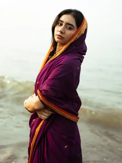 The deep purple of the saree against the muted tones of the sea creates a rich, moody, and almost painterly portrait.