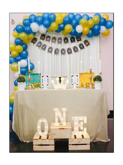 A royal celebration for a little prince's first birthday. We designed this dessert table with a blue, yellow, and white balloon arch, a 'Happy Birthday' banner, and light-up marquee letters spelling 'ONE' on rustic wooden crates.