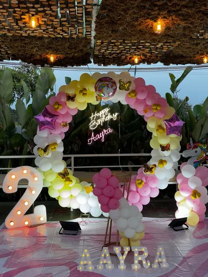 A unicorn-themed birthday setup with a circular balloon arch in pastel rainbow colors. The design is accented with butterfly and star-shaped foil balloons, a neon sign with the child's name, and a large light-up number '2'.