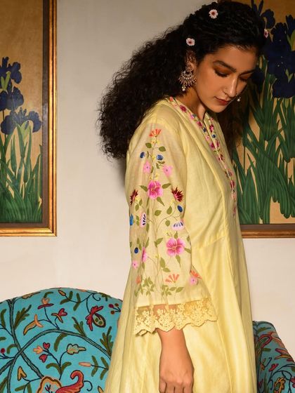 A close-up on the delicate floral embroidery of a yellow kurta from Baavli. This shot emphasizes the craftsmanship and artistry of the garment, set against a backdrop of floral paintings that echo the design.