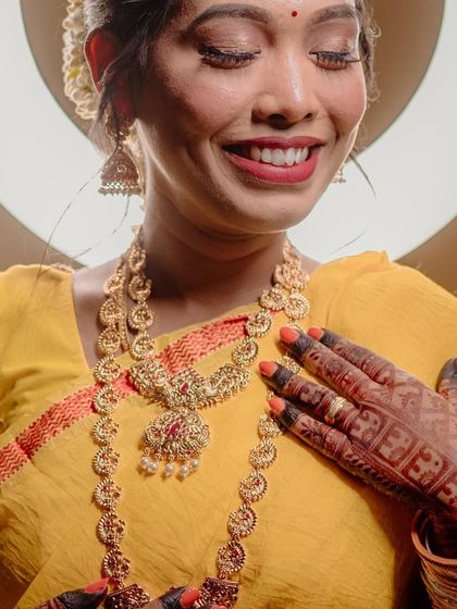 A close-up portrait of a South Indian bride, her hand resting on her chest, drawing attention to her beautiful henna and traditional gold jewelry.