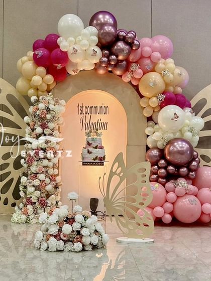 An elegant butterfly-themed stage for a First Communion, decorated in shades of pink and mauve. The setup includes a beautiful balloon arch, floral towers, and a custom backdrop with the child's name.
