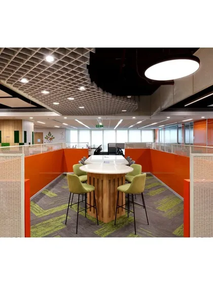 A circular high-top meeting table nestled within a low-partitioned area, offering a space for informal collaboration. The design uses a mix of textures, from the wooden base to the patterned carpet and acoustic ceiling panels.