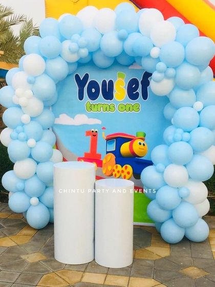 A Thomas and Friends themed first birthday party. The outdoor setup includes a blue and white balloon arch framing a backdrop with the beloved train character.