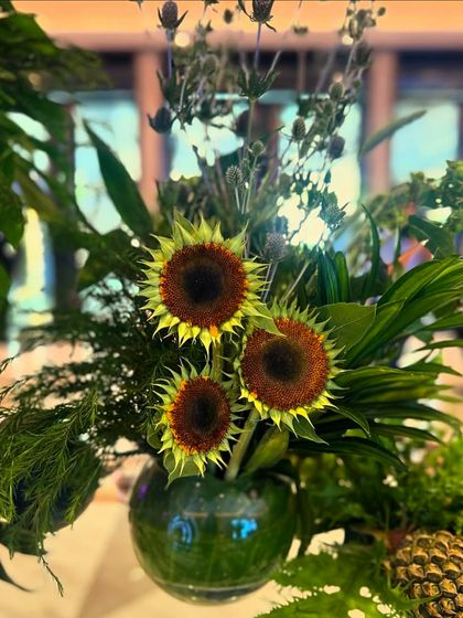 A close-up of a simple vase of sunflowers, a cheerful detail amidst the larger, more complex foliage arrangements on the dessert table.