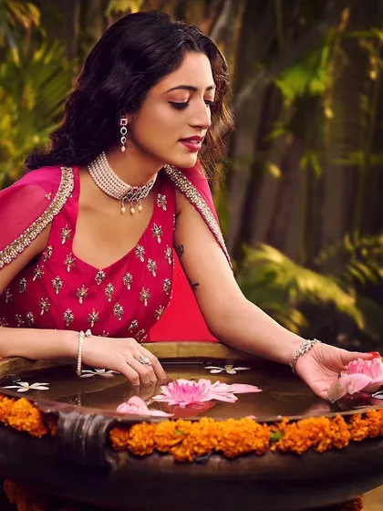 A serene moment from a festive jewellery campaign. The model, dressed in a vibrant pink outfit, interacts with lotus flowers in an urli, creating a peaceful and elegant scene that highlights the pearl choker and earrings.