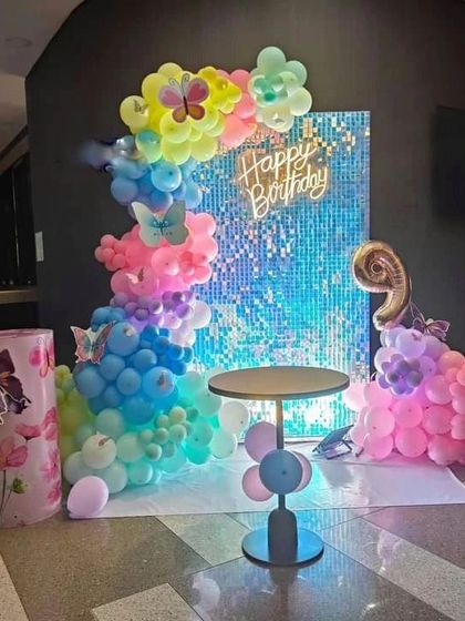 A colorful butterfly-themed decoration against a blue sequin wall. The setup features a rainbow balloon garland, a 'Happy Birthday' neon sign, and a themed pedestal.