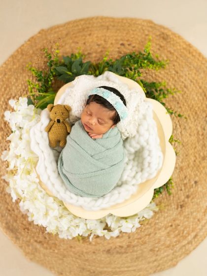 An overhead shot of a swaddled newborn in a flower-shaped basket, placed on a rustic jute mat with a tiny teddy bear prop.