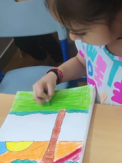 A close-up of a student coloring a landscape. This helps them learn about different elements like the sun, trees, and ground.