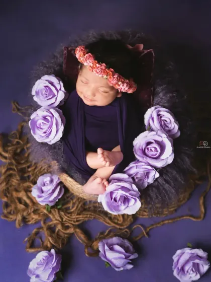 A sleeping newborn is nestled in a basket, surrounded by beautiful purple roses against a rich purple backdrop. The floral crown adds a final touch to this artistic composition.