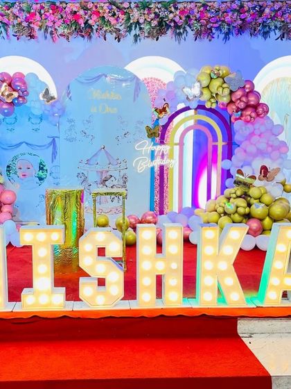 An elaborate and colorful stage decoration for a first birthday. This setup features multiple arched backdrops, a floral ceiling, and large light-up letters spelling "MISHKA".