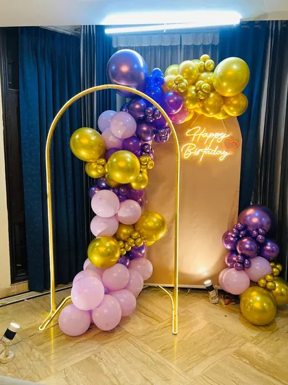 A birthday backdrop featuring a golden metal arch adorned with a garland of purple, pink, and gold balloons and a "Happy Birthday" neon sign.