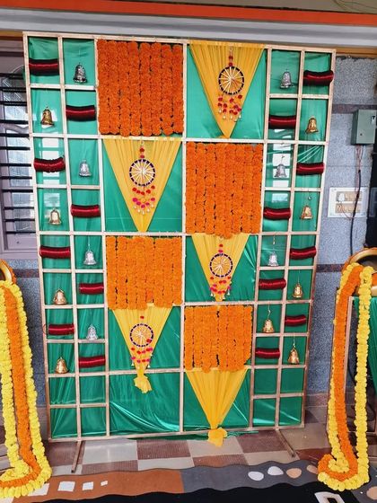 A traditional backdrop for a Bangle ceremony (Balesh Shastra). The design uses a grid pattern with panels of marigolds, fabric, and hanging bells, with decorative pots of overflowing flowers.