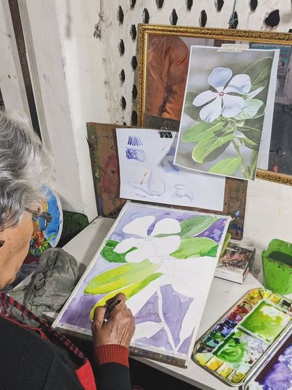An overhead view of an artist at work. Harinder ma'am is carefully painting a white flower, demonstrating her delicate touch with watercolors.