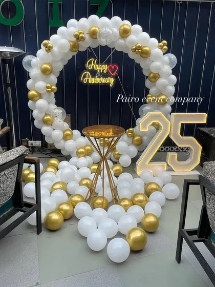 An elegant 25th-anniversary party backdrop. This features a full circle balloon arch in a sophisticated white and gold color scheme, a "Happy Anniversary" neon sign, and large "25" marquee numbers.
