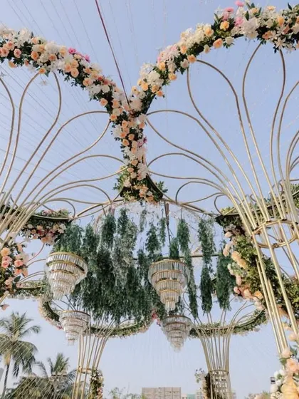A low-angle shot of the heart-shaped floral mandap, showing the intricate structure and the chandeliers hanging within, against a clear blue sky.