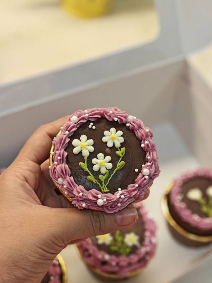 Holding up one of my floral cupcakes. This design features delicate, hand-piped daisies on a rich chocolate ganache base, framed by a purple buttercream border.