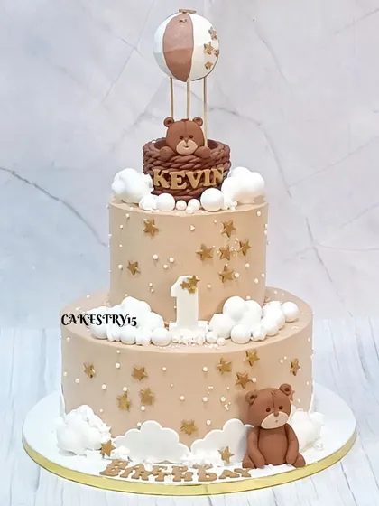 A whimsical two-tier coffee caramel cake for a first birthday. The design features a teddy bear in a hot air balloon, surrounded by fondant clouds and golden stars.
