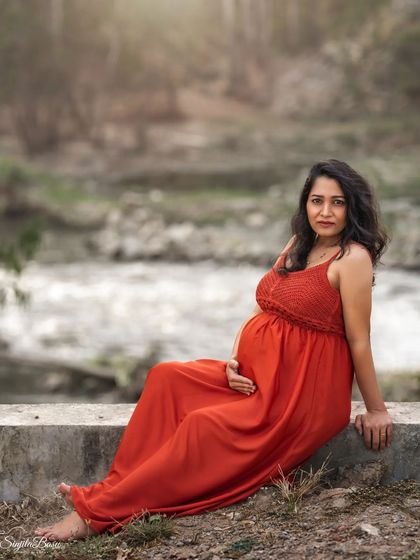 A duplicate of ID 1, this outdoor portrait shows an expecting mother in a flowing red dress sitting peacefully by a flowing stream, combining the beauty of nature with a serene mood.