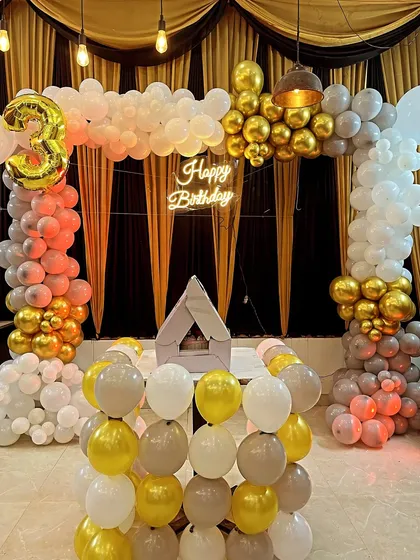 A stylish birthday setup with a pastel and gold balloon arch. The decoration includes a neon sign and a small house-shaped prop.