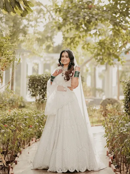 A full-length shot of the bride in a beautiful garden path. Her all-white lehenga stands out beautifully against the lush greenery, creating a fresh and elegant bridal portrait.