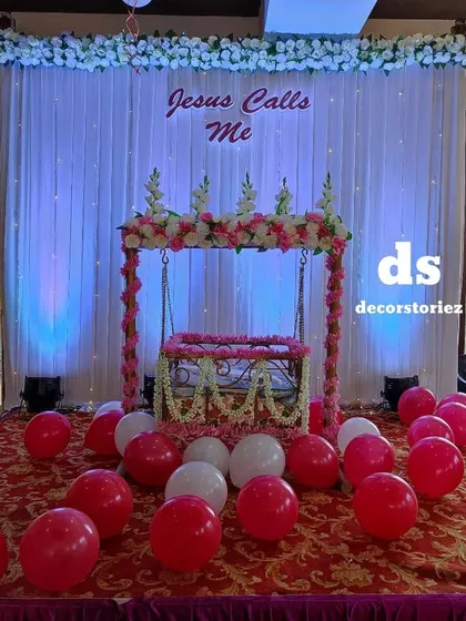 A wider angle of the naming ceremony setup, showing the decorated cradle on a red carpeted stage. The backdrop is topped with a white floral border, tying the whole look together.