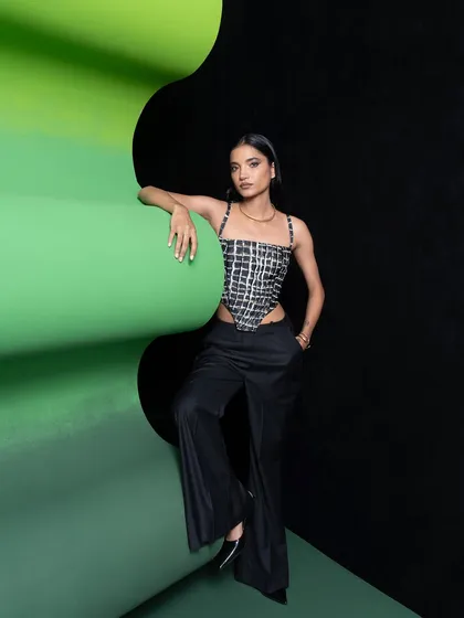 A full-length shot of a single model, showcasing a modern look with a grid-patterned corset and wide-leg trousers. The pose is relaxed yet confident, set against the graphic backdrop.