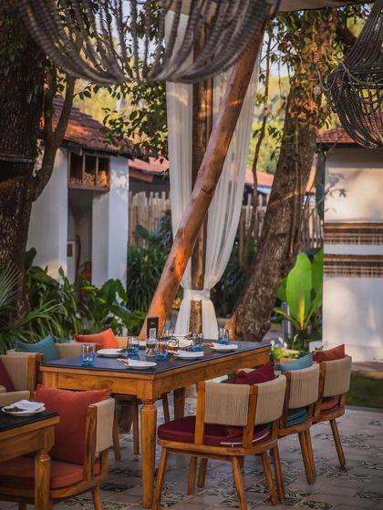 An outdoor dining area at Sanctuary Bar in Goa, featured in Architectural Digest. The setting combines rustic wooden furniture with colorful cushions, all under a canopy surrounded by tropical greenery.