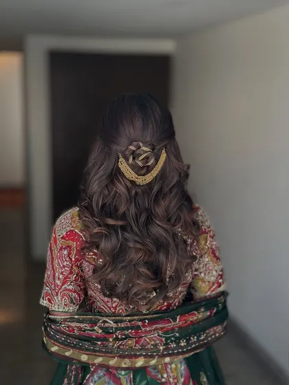 A beautiful half-updo for a Mehndi ceremony, featuring a rose-shaped braid and a golden accessory. The rest of the hair is styled in soft waves.