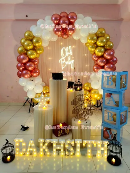 An elegant 'Oh Baby' setup with a balloon arch in rose gold, gold, and white. The decor is complete with pedestals, marquee letters, and 'BABY' blocks.