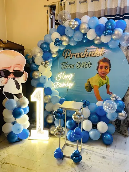 A personalized Boss Baby first birthday backdrop. The setup includes a large circular print with the baby's photo, a character cutout, and a blue, white, and silver balloon arch.