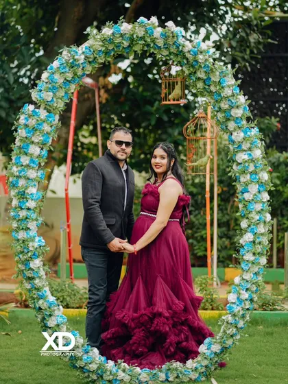 A formal portrait of a couple framed by a large floral arch, looking directly at the camera.