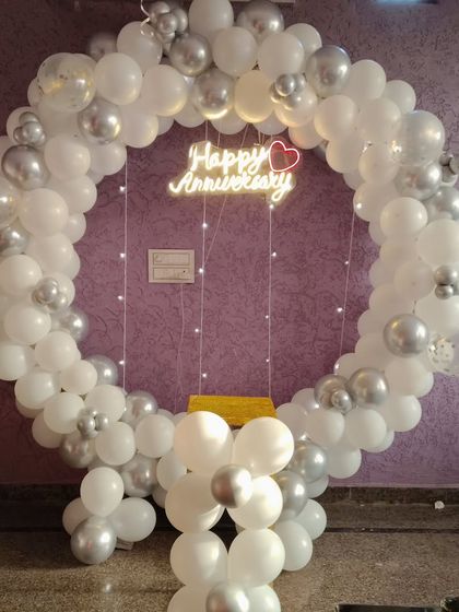 For a simple yet beautiful at-home anniversary surprise, I can set up a clean and classic balloon ring. This one uses white and silver balloons with a "Happy Anniversary" neon sign, creating a lovely backdrop for your celebration.