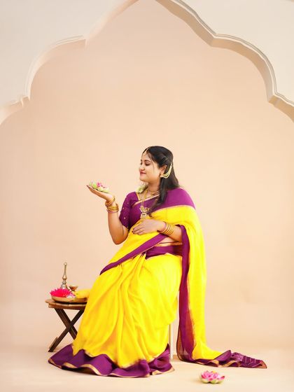 A graceful solo portrait of a mother-to-be in a yellow and purple saree, seated elegantly in a traditional studio setup.