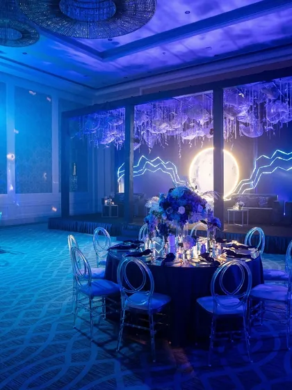 A stunning dining setup for a "moonlit" themed event. The entire room is bathed in blue light, with a large illuminated moon as the stage backdrop.