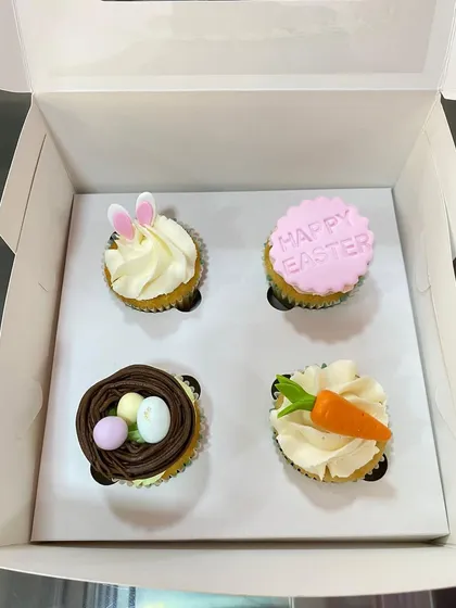 A box of four assorted Easter-themed cupcakes, featuring bunny ears, a bird's nest, a carrot, and a "Happy Easter" message.