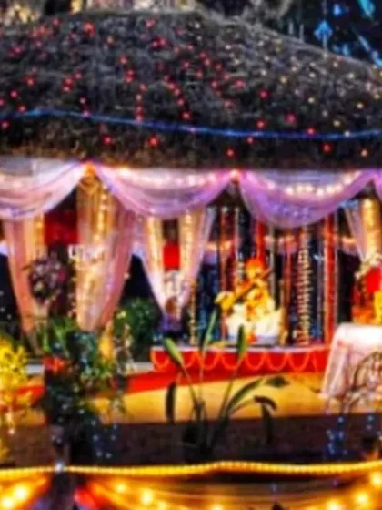 An outdoor Ganpati setup inside a gazebo structure. The hut-like roof and surrounding area are decorated with colorful string lights, creating a warm and inviting atmosphere for evening darshan.