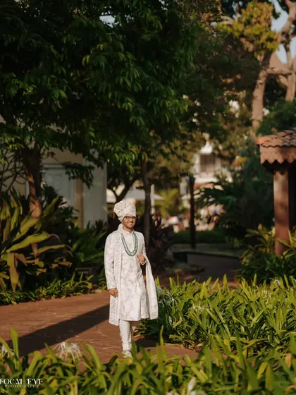 A full-length portrait of the groom in his elegant white and green sherwani, standing confidently in a lush garden setting.