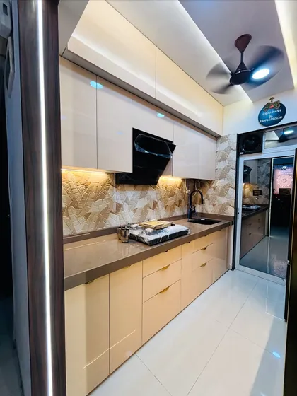 This parallel kitchen design creates a warm and luxurious feel with high-gloss beige cabinets and ambient lighting. Note the textured stone backsplash and integrated black chimney, creating a seamless and elegant workspace.