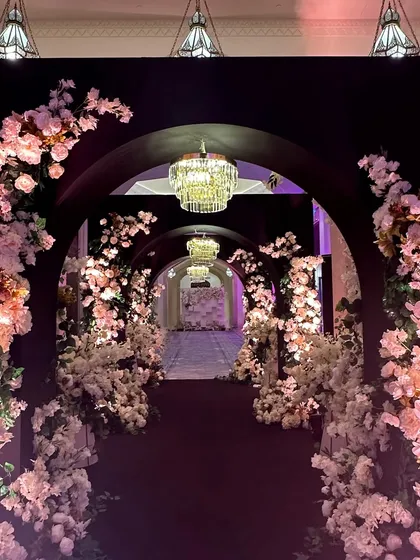 A journey through a tunnel of elegance. This walkway, framed by dark arches and cascading white and pink blossoms, leads guests into a magical reception space.