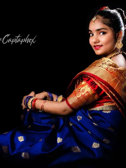 A three-quarter seated portrait against a black background. The pose looking back over the shoulder is classic and graceful, highlighting the rich border of the saree and traditional jewelry.