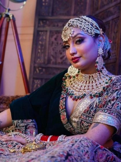 A bride seated in a dark green and red lehenga with intricate embroidery, paired with a heavy multi-colored jewellery set.